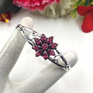 May include: A silver-toned metal cuff bracelet featuring a central flower-shaped ornament. The flower is composed of marquise-cut, deep pink faceted crystals arranged in a radial pattern around a single round, central pink crystal stone.