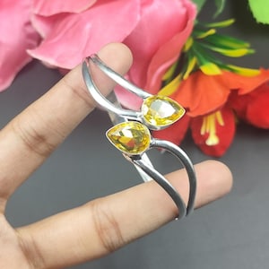 May include: A silver bracelet with two yellow, teardrop-shaped gemstones. The bracelet curves around the wrist. The background features pink and orange flowers. The gemstones are set at opposite ends.