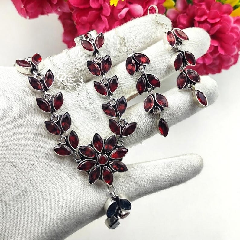 May include: A jewellery set consisting of a necklace and matching drop earrings, crafted from silver-toned metal and featuring marquise-cut deep red garnet stones arranged in leaf-like patterns, with a central flower-shaped pendant on the necklace.