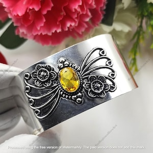 May include: A wide silver-toned cuff bracelet featuring a central oval-cut yellow gemstone. The metal surface is decorated with embossed floral motifs and intricate swirling filigree patterns that extend outward from the central stone setting.