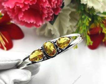 May include: A silver bracelet with three yellow gemstones, each approximately 1.5 cm in length. The bracelet has a decorative setting and a polished finish. The background features vibrant flowers.