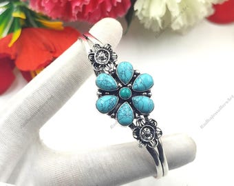 May include: A silver-toned cuff bracelet featuring a central flower motif composed of six teardrop-shaped turquoise stones with dark veining and a small round turquoise centre, flanked by two smaller silver floral accents on the band.