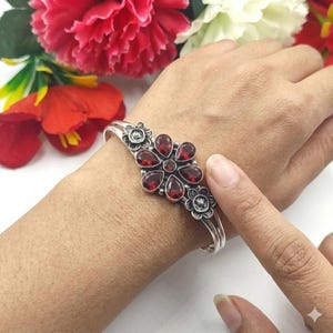 Può includere: Un bracciale rigido in argento con un motivo floreale centrale composto da sei pietre preziose granato rosso scuro a forma di pera che circondano un piccolo granato rotondo centrale, affiancato da due piccoli accenti floreali in argento con petali testurizzati.
