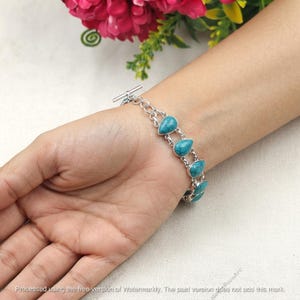 May include: A silver-toned chain bracelet featuring multiple teardrop-shaped turquoise stones set in metal bezels. The bracelet is secured with a toggle clasp mechanism, showcasing a series of linked segments that wrap around the wrist area.
