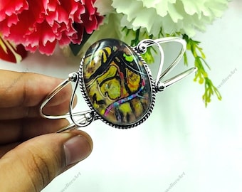 May include: Silver cuff bracelet with an oval stone displaying an abstract pattern in yellow, black, and purple. The stone is set in a textured bezel. The bracelet has a simple, open design.