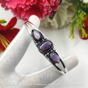Peut inclure: Un bracelet argenté orné de trois pierres précieuses améthystes violettes en forme de goutte. Le bracelet est de type jonc ouvert. Le fond présente des fleurs roses et rouges.