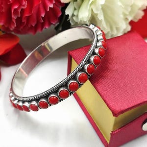 May include: A silver bangle bracelet featuring a row of round, vibrant red stones set along the outer edge. The metal band has a polished finish and a twisted rope-like texture bordering the stones, creating a detailed and structured appearance.