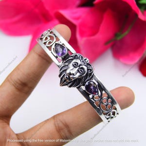 May include: Silver-toned bracelet featuring a lion's head centerpiece and two purple gemstones. The bracelet has an openwork design with decorative swirls. The gemstones are teardrop and round shaped.