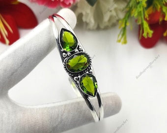 May include: A silver cuff bracelet featuring three faceted green peridot gemstones. The centre stone is oval-shaped, flanked by two pear-shaped stones. The metal setting displays a twisted rope texture around the centre stone and smooth bands.