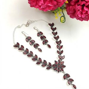 May include: A silver-toned necklace and matching pair of drop earrings featuring marquise-cut deep red stones arranged in a leaf-like pattern. The necklace includes a central floral motif with a dangling segment, all connected by a delicate silver chain.