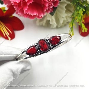 May include: A silver bracelet with three bright red gemstones. The gemstones are oval and teardrop-shaped, set in a polished metal band. The bracelet is shown against a backdrop of pink and red flowers.