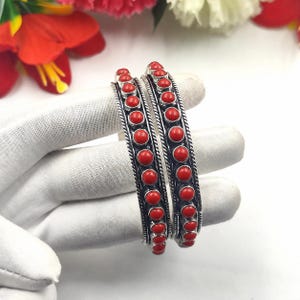 Peut inclure: Deux bracelets joncs en argent ornés d'une rangée centrale de pierres rondes rouge vif. Chaque bracelet est encadré par des détails en fil d'argent torsadé sur les deux côtés, créant un aspect texturé. Les pierres sont disposées en ligne.