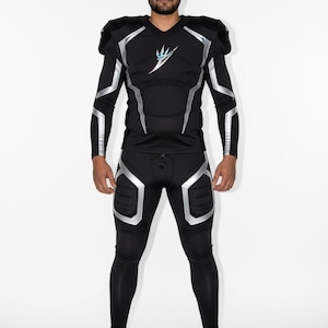 Op de afbeelding: Zwart voetbaluniform met zilveren accenten. Het uniform bestaat uit een shirt met lange mouwen en schouderbeschermers, leggings en kniebeschermers. Een logo is zichtbaar op de borst. De persoon draagt zwarte voetbalschoenen.