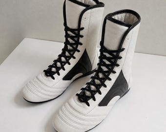May include: A pair of high-top boxing boots featuring a white textured leather upper with contrasting black leather side panels and black laces. The boots have a rounded toe, a high ankle collar, and a flat sole, designed for athletic performance.