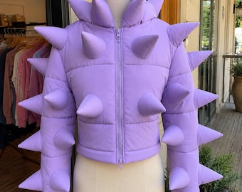 May include: A light purple puffer jacket featuring a high collar and numerous padded, cone-shaped spikes extending from the shoulders, chest, and sleeves. The jacket has a central vertical zipper closure and a cropped, fitted silhouette.