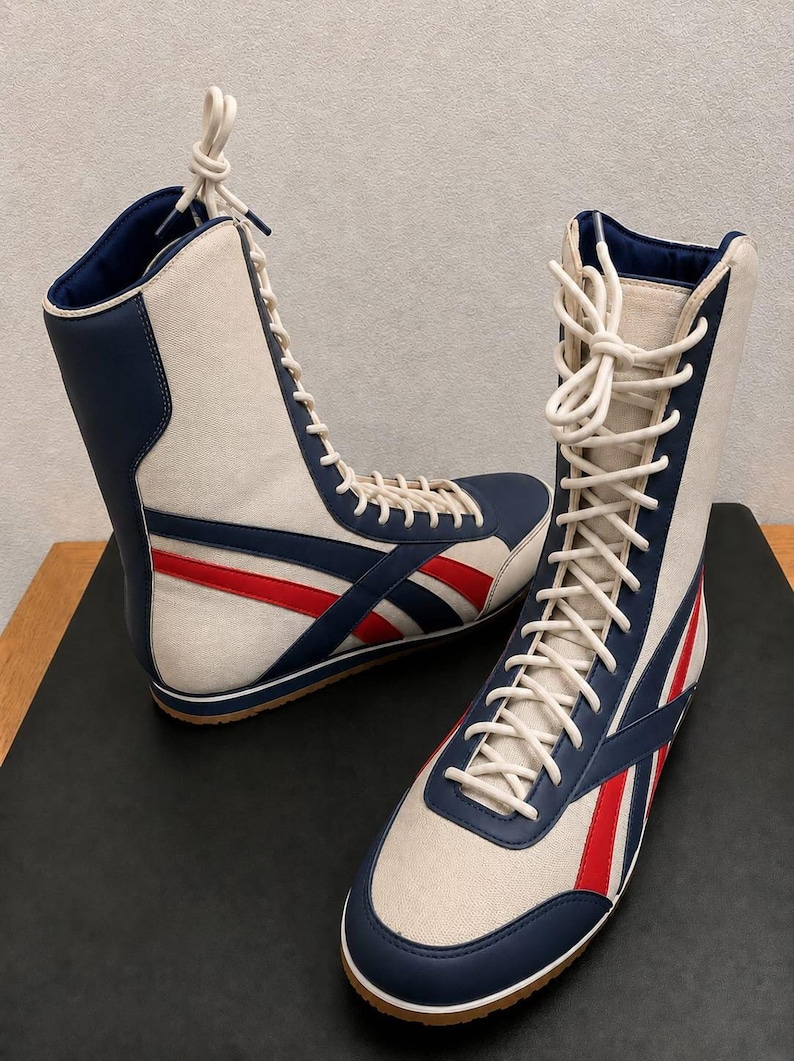 A pair of high-top white leather wrestling boots with blue and red side stripes, white laces tied at the top, and gum rubber soles, standing on a black surface.