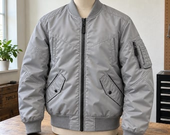 May include: A light gray bomber jacket featuring a full-length black front zipper, a ribbed collar, cuffs, and hem. The jacket includes two front pockets with snap closures, a zippered utility pocket on the left sleeve, and decorative seams.