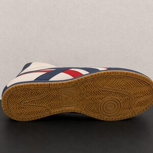 Underside view of a wrestling boot displaying the brown gum rubber traction sole with a textured geometric pattern and a central letter logo.