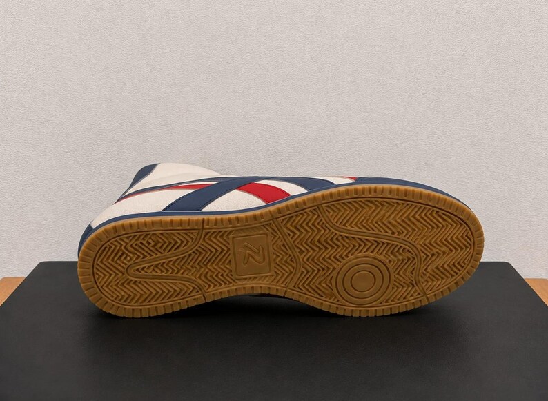Underside view of a wrestling boot displaying the brown gum rubber traction sole with a textured geometric pattern and a central letter logo.
