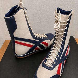 Pair of high-top white leather boots with blue and red athletic stripes, white laces, and gum soles, displayed on a dark platform.