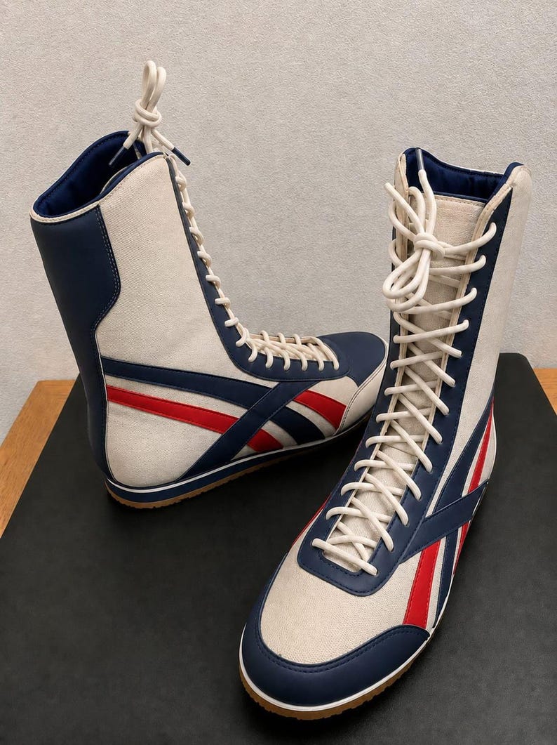 Pair of high-top white leather boots with blue and red athletic stripes, white laces, and gum soles, displayed on a dark platform.