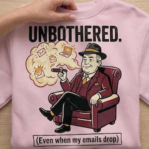 May include: Pink sweatshirt with the text "UNBOTHERED." and a cartoon illustration of a man in a suit smoking a cigar, seated in a red armchair. The image includes a thought bubble with email and laptop graphics, and the text "(Even when my emails drop)".