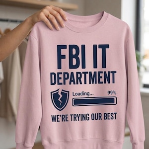 Könnte beinhalten: Ein hellrosa Sweatshirt mit dem marineblauen Text "FBI IT DEPARTMENT". Darunter befindet sich eine Grafik eines Schildes mit einem Blitz und einer Ladeleiste, die "99%" anzeigt. Der Satz "WE'RE TRYING OUR BEST" ist darunter in Marineblau gedruckt.