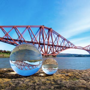 May include: A scenic photograph featuring the Forth Bridge in Scotland, reflected in two glass spheres. The bridge is a vibrant red, set against a clear blue sky and water. The spheres sit on a textured, brown surface.