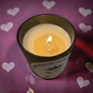May include: Lit candle in a black glass container with the words "I Love You!" written on the wax. The candle is on a purple surface with white heart shapes.