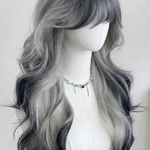 Peut inclure: Une longue perruque ondulée avec un mélange de gris et de noir, coiffée d'une frange. La perruque est présentée sur une tête de mannequin, mettant en valeur son design en couches et son dégradé de couleurs. Un collier argenté avec des breloques est également visible.