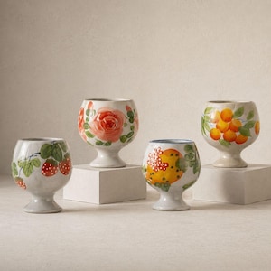 May include: Four white ceramic goblets with hand-painted fruit and floral designs. The designs include strawberries, a rose, oranges, and a yellow fruit. Each goblet is on a small, white square pedestal.