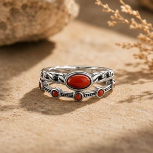May include: Two silver rings with red gemstones. The top ring has a chain-like design and an oval red stone. The bottom ring features small, round red stones. The rings are displayed on a tan surface, with a natural background.