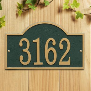 Puede incluir: Placa de dirección verde oscuro con números y bordes dorados. Los números "3162" se muestran en la placa. La placa es rectangular con una parte superior arqueada y está montada sobre una superficie de madera.