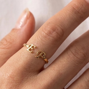 May include: A gold-coloured open ring featuring the letters "E" and "P". The ring is worn on a finger. The ring is made of a shiny metal and is set against a soft, neutral background.