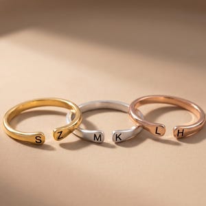 May include: Three open-ended rings in gold, silver, and rose gold. Each ring features a single initial: S, Z, M, K, L, and H. The rings are displayed on a neutral background.