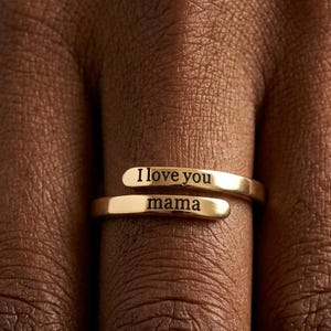 Könnte beinhalten: Ein goldfarbener Wickelring mit zwei überlappenden Bändern. Das obere Band ist mit dem Text I love you graviert, das untere Band mit dem Text mama. Der Ring hat eine polierte metallische Oberfläche und ein offenes, verstellbares Design.