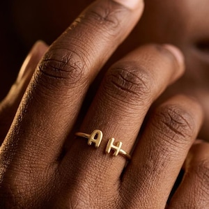 May include: A gold-toned open ring featuring two capital letters, A and H, positioned at the ends of the band. The ring has a polished metallic finish and a simple, thin band design that remains open between the two letters.