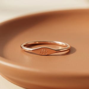 May include: A rose gold ring featuring a thin, polished band and a small, centered, slightly elongated oval plate. The plate is engraved with the lowercase cursive letter m. The ring has a smooth, metallic finish and a consistent rose gold tone.