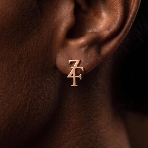 May include: A rose gold stud earring featuring the stylized letters Z and F intertwined. The design has a polished metallic finish and is positioned on an earlobe. The letter Z sits above the letter F, creating a singular, cohesive monogram shape.