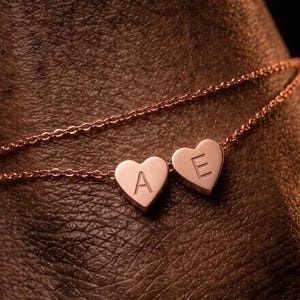 Peut inclure: Un bracelet à chaîne de couleur or rose orné de deux petits pendentifs en forme de cœur. Chaque cœur est gravé d'une lettre majuscule, respectivement A et E, centrée sur la surface lisse et polie du métal de chaque breloque.