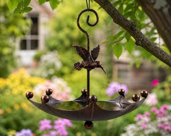 May include: A bronze-colored hummingbird feeder hanging from a tree branch. The feeder has an umbrella-shaped dish with decorative spheres and a hummingbird figurine perched on a central post. The background features a garden with colorful flowers.