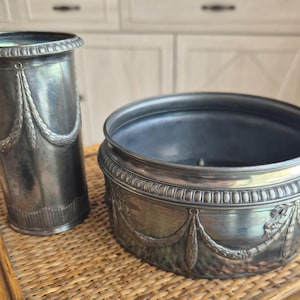 Pode incluir: Um vaso cilíndrico alto de tom prateado e uma taça circular larga de tom prateado. Ambos os itens apresentam um padrão decorativo em relevo a condizer de festões drapeados e caneluras verticais na base, com um detalhe de rebordo perlado.