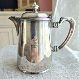 Peut inclure: Une cafetière argentée avec un couvercle et une poignée. La cafetière a un bec verseur et un petit emblème près de la base. La poignée a un design décoratif. La cafetière est posée sur une surface claire.