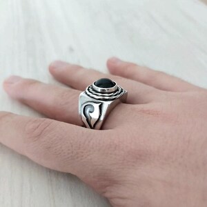 May include: A silver ring featuring a smooth, round, black stone set in the center. The stone is surrounded by a twisted metal wire design. The sides of the ring band are wide and decorated with a carved, swirling, wave-like pattern in the metal.