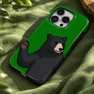 May include: A green smartphone case featuring a cartoon illustration of a black bear with a tan muzzle waving its paw. The case has a cutout for a triple-lens camera system on the back and a black border surrounding the edges of the device.