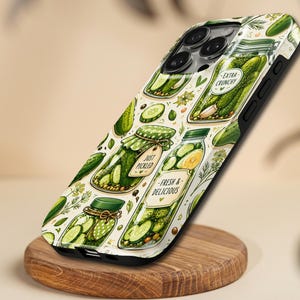 Op de afbeelding: Een telefoonhoesje met een herhalend patroon van cartoonpotten gevuld met augurken. De potten hebben groene deksels en labels met de tekst "Extra Crunchy", "Just Pickled" en "Fresh & Delicious". De achtergrond is lichtcrème van kleur.