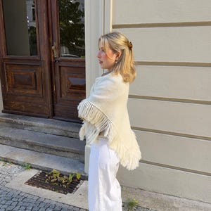 Può includere: Un poncho in maglia color crema con una trama testurizzata e lunghe frange lungo i bordi, abbinato a pantaloni bianchi a gamba larga e scarpe di colore chiaro. Il capo è indossato sopra un top di colore neutro, creando un look a strati.
