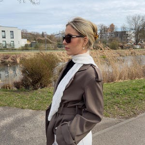 Könnte beinhalten: Ein brauner Trenchcoat mit Gürteltaille und Schulterklappen, getragen über einem schwarzen Oberteil und einer weißen Hose. Ein schmaler, weißer, strukturierter Schal liegt um den Hals. Das Outfit umfasst eine dunkle Sonnenbrille und eine Haarspange.