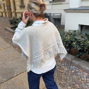Könnte beinhalten: Ein beigefarbener Strickponcho mit einer strukturierten Zopfmusterborte und langen Fransen am Saum, getragen über einem weißen Langarmshirt und einer dunkelblauen Jeans. Das Kleidungsstück hat eine weite, drapierte Silhouette mit weicher Textur.