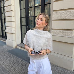 Könnte beinhalten: Eine Frau trägt einen hellgrauen, kurzärmeligen Pullover und weiße, plissierte Shorts. Sie hält eine schwarze Sonnenbrille. Der Hintergrund zeigt ein Gebäude mit großen Fenstern und einer Steinfassade. Das Outfit ist für warmes Wetter geeignet.